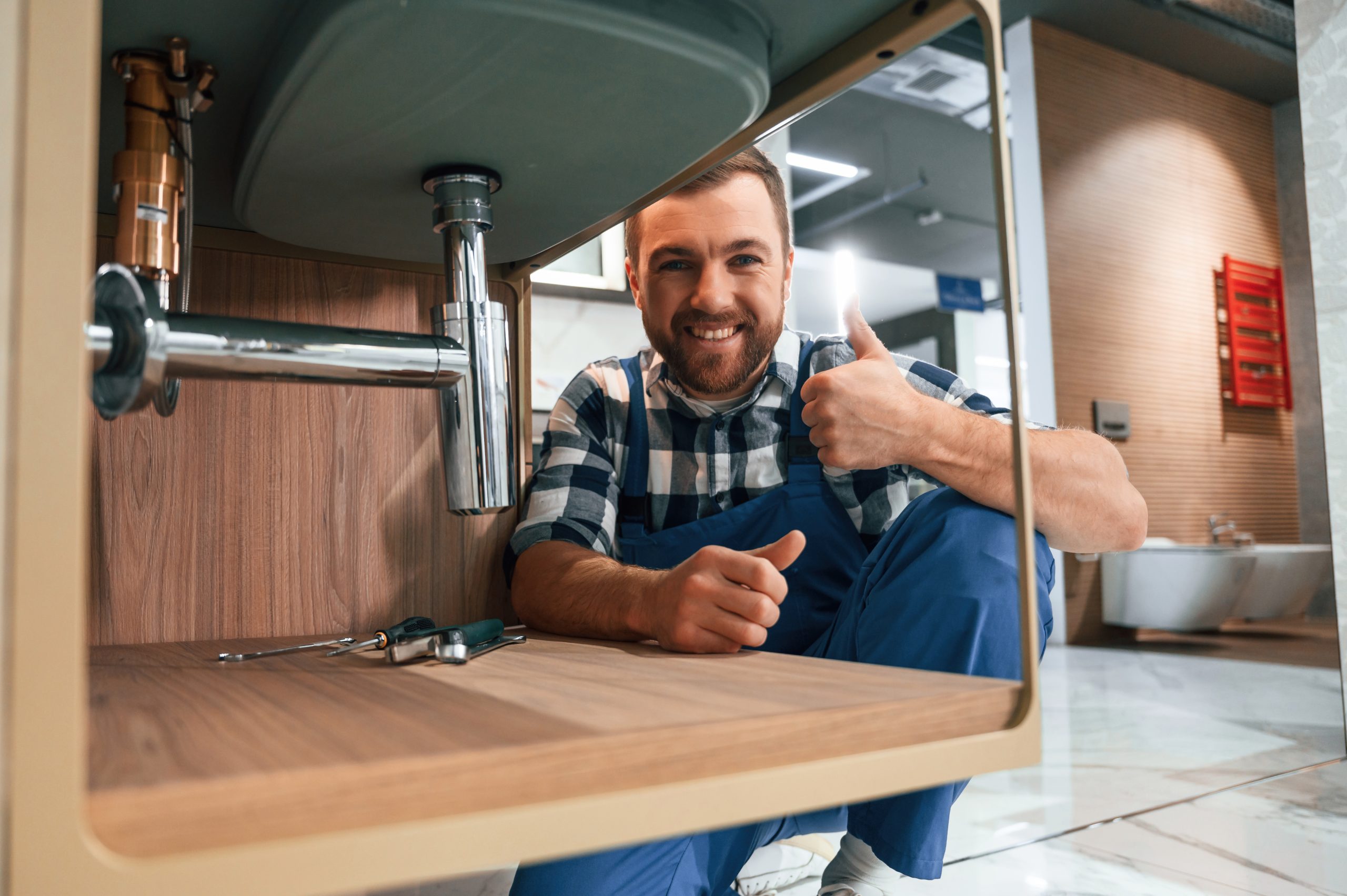 Shwoing thumb up and smiling. Plumber in blue uniform is at work in the bathroom